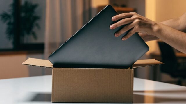 Hands lifting a brand-new graphite black laptop out of its plain cardboard box on a white desk