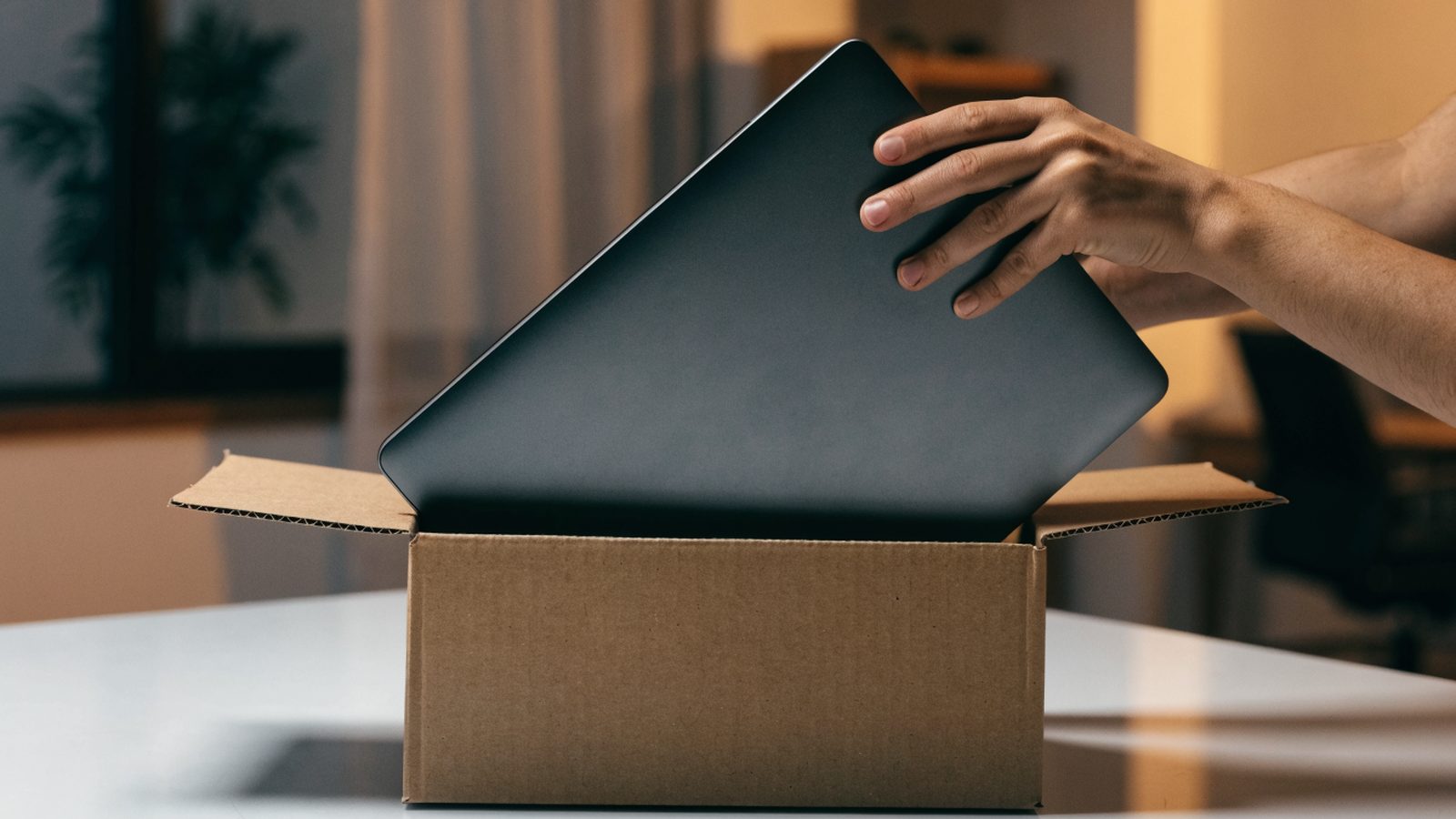Hands lifting a brand-new graphite black laptop out of its plain cardboard box on a white desk