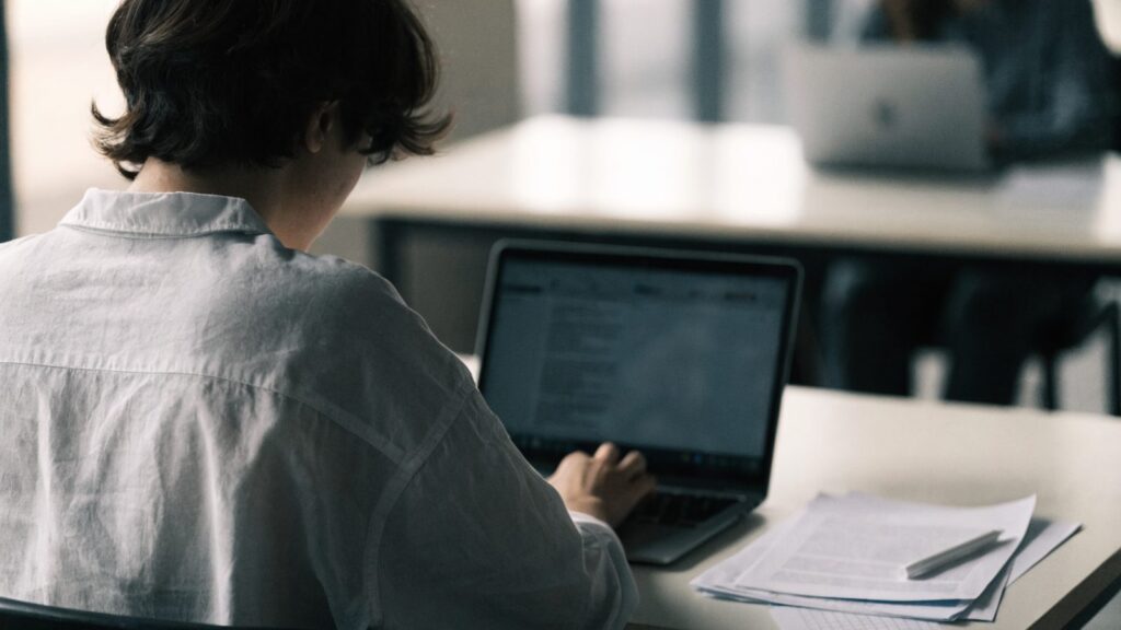A teenager seen from behind doing homework on a matte grey laptop at a desk