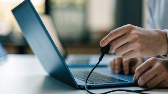 Hands unplugging a charging cable from a slim dark blue laptop on a white desk