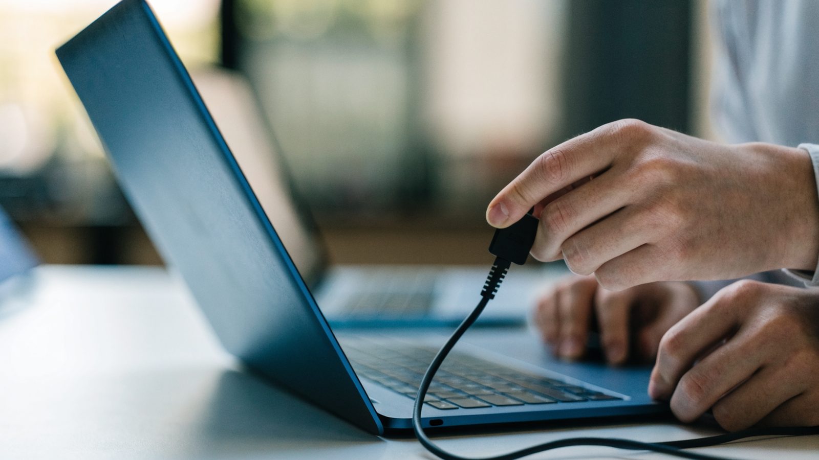 Hands unplugging a charging cable from a slim dark blue laptop on a white desk