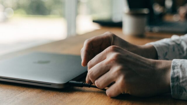 Hands unplugging a charging cable from a slim matte grey laptop on a wooden desk