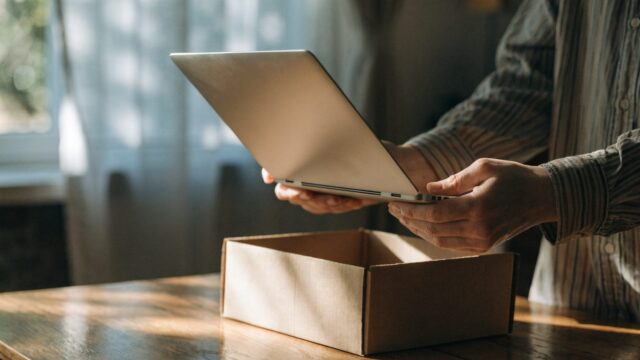 Hands lifting a brand-new silver laptop out of its plain cardboard box on a wooden desk