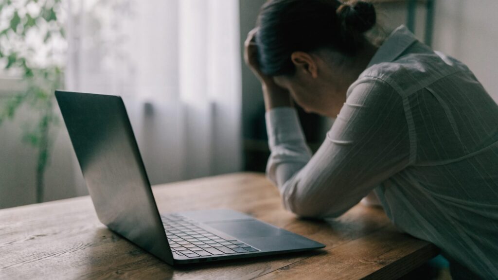 A person resting their forehead on one hand in front of a graphite black laptop on a wooden desk