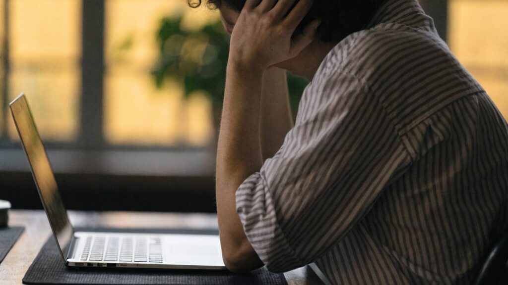 A person resting their forehead on one hand in front of a silver laptop on a dark desk mat
