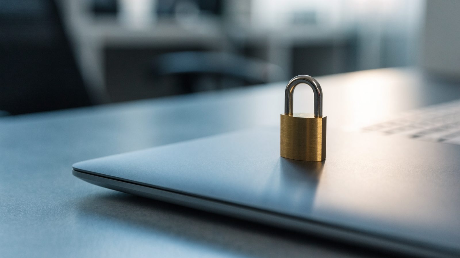A small brass padlock resting on the closed lid of a matte grey laptop on an office desk