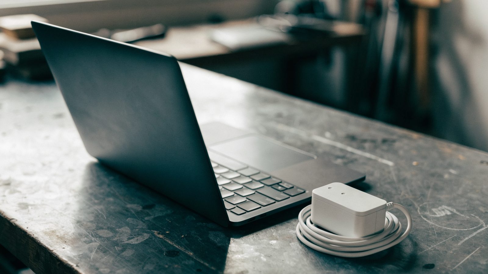 A graphite black laptop on a workbench next to a compact white power adapter with its cable neatly coiled