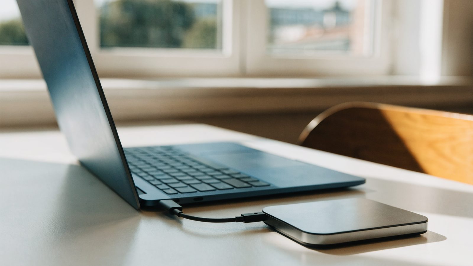 A small external hard drive connected by cable to a dark blue laptop on a white desk