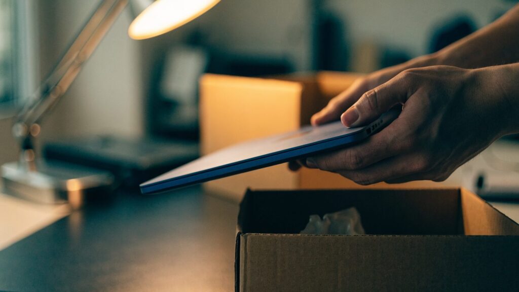 Hands lifting a brand-new dark blue laptop out of its plain cardboard box on a dark desk mat