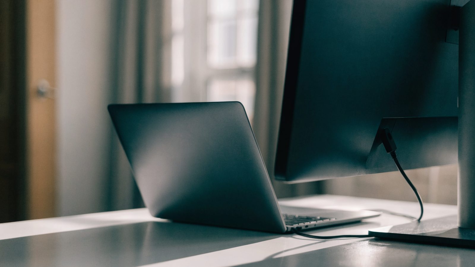 A graphite black laptop connected to a large external monitor with a blank dark screen via a cable on a white