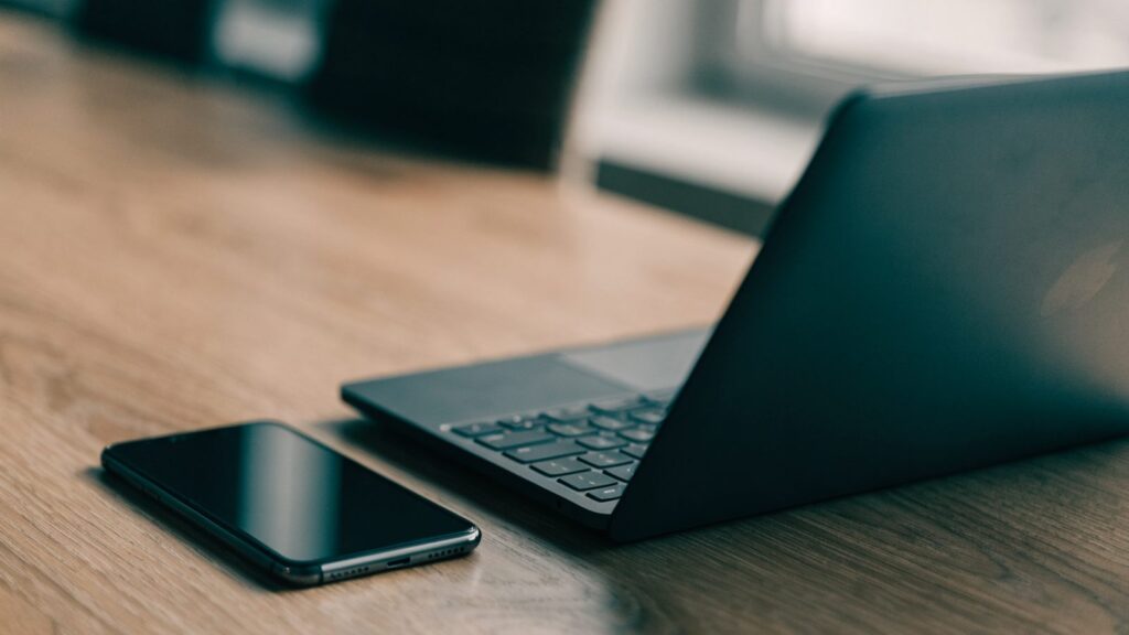 A smartphone lying beside a graphite black laptop on a wooden desk