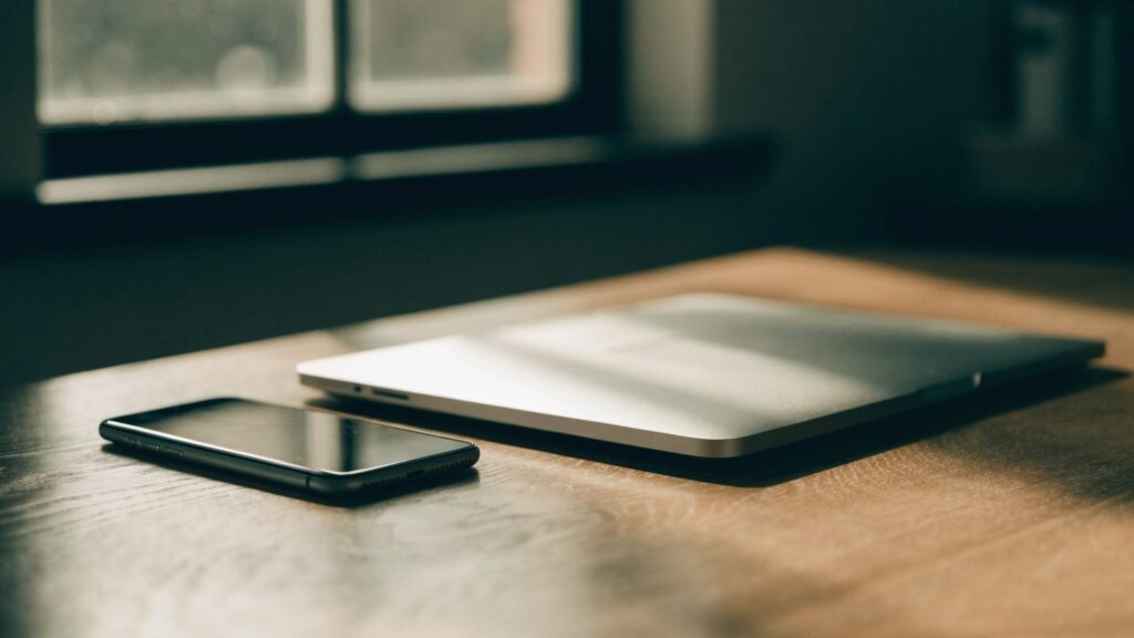 A smartphone lying beside a silver laptop on a wooden desk