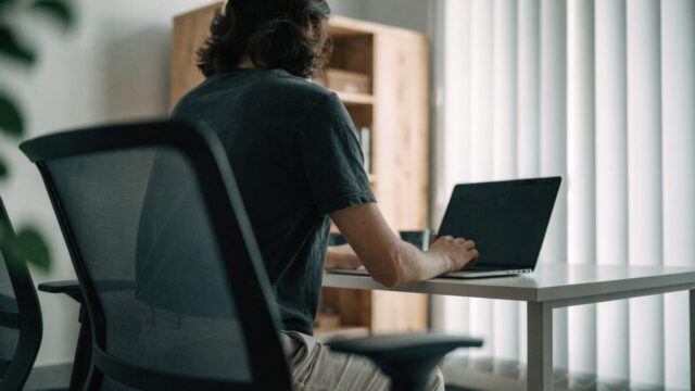 A person seen from behind working on a matte grey laptop in a bright home office