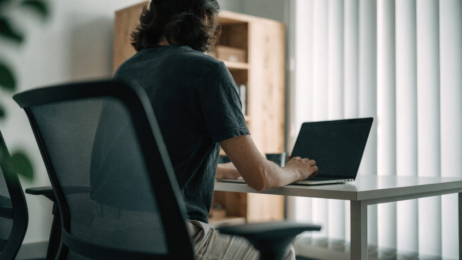 A person seen from behind working on a matte grey laptop in a bright home office