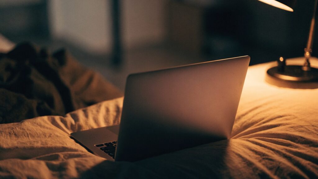 A matte grey laptop glowing softly on a bed in a dark room