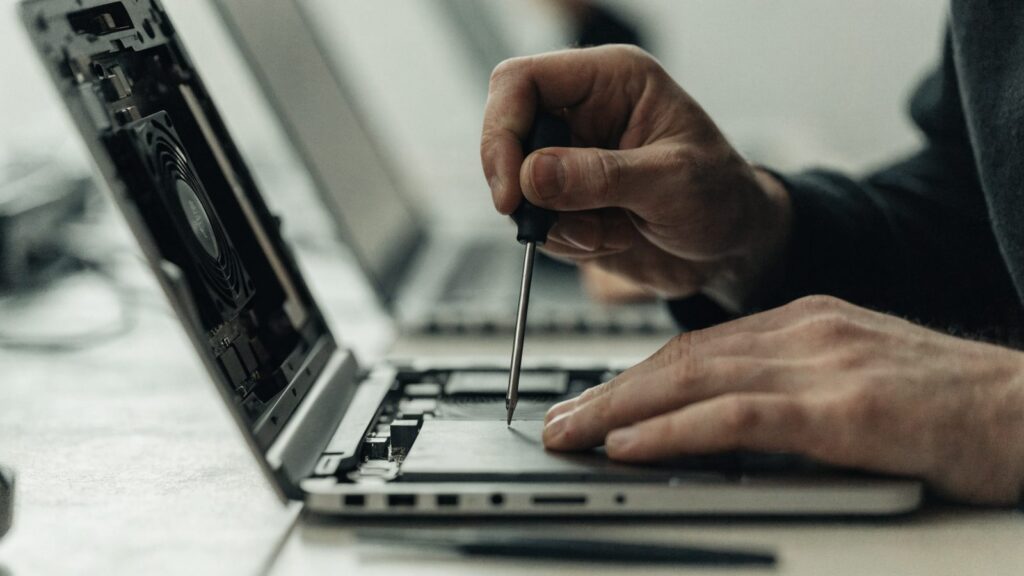 A technician's hands holding a small screwdriver over an opened silver laptop with internal components visible