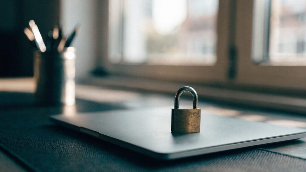 A small brass padlock resting on the closed lid of a matte grey laptop on a dark desk mat