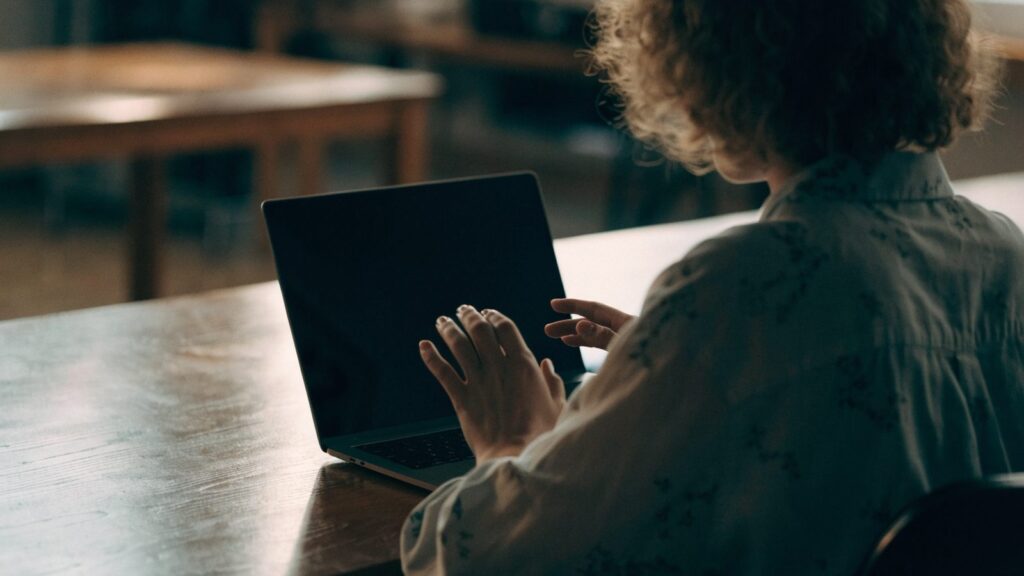 A person seen from behind with hands raised at a matte grey laptop with a dark blank screen on a wooden desk