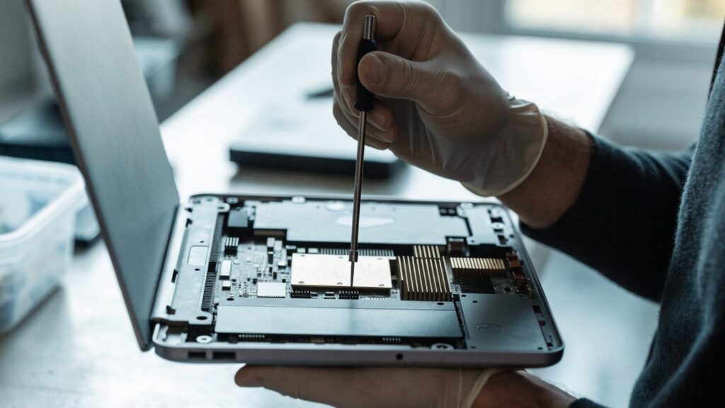 A technician's hands holding a small screwdriver over an opened matte grey laptop with internal components vis