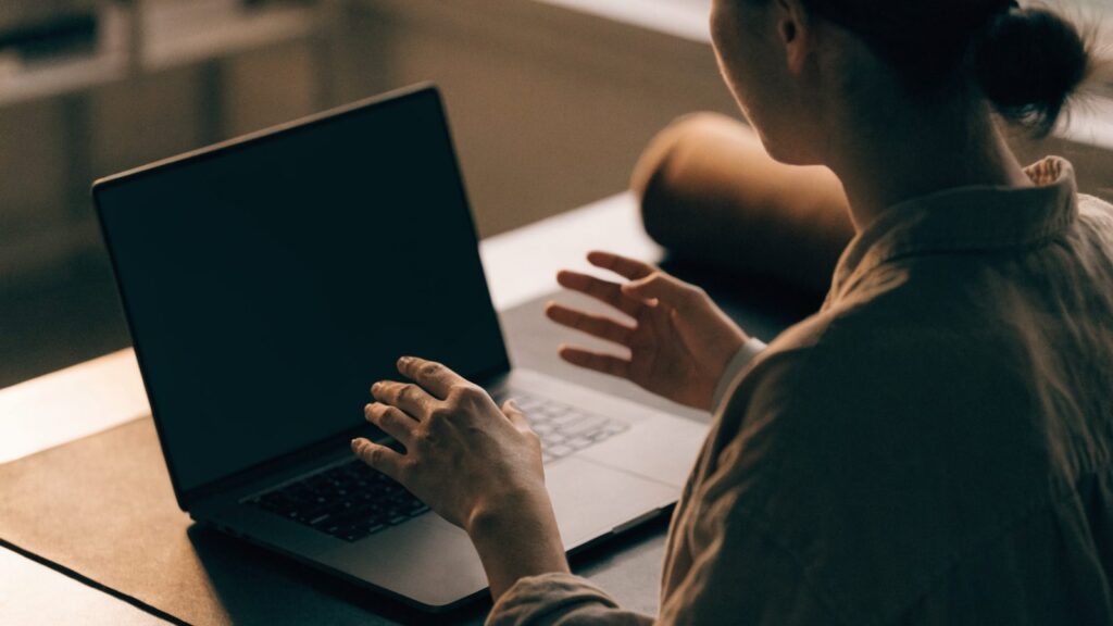 A person seen from behind with hands raised at a graphite black laptop with a dark blank screen on a dark desk