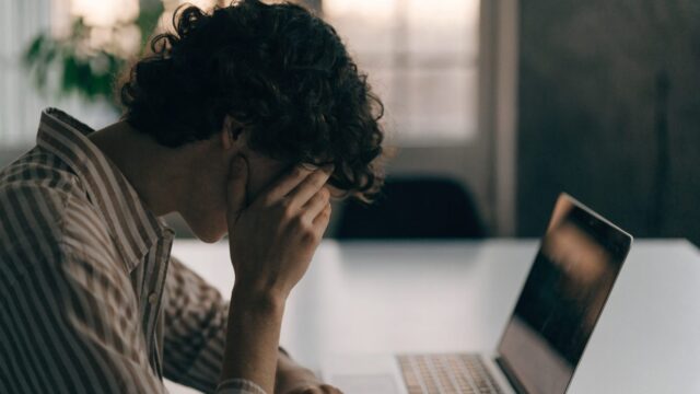 A person resting their forehead on one hand in front of a silver laptop on a white desk