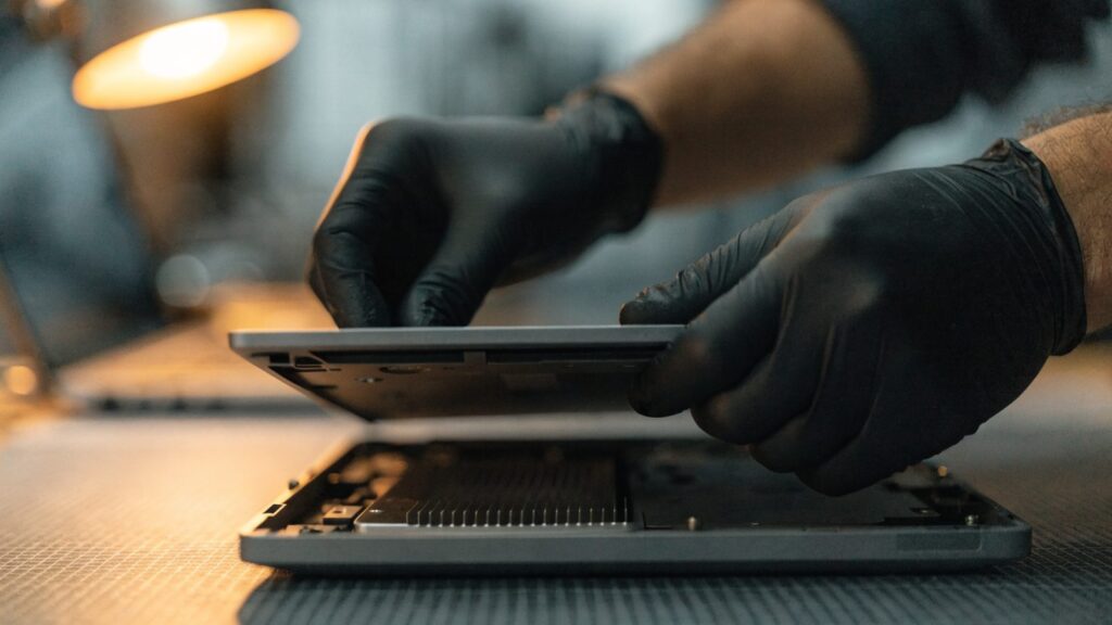 A technician lifting the bottom cover off a matte grey laptop on an anti-static mat