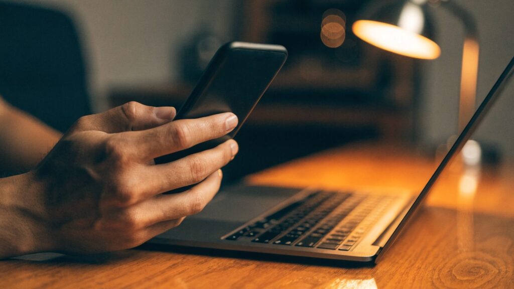 A hand holding a smartphone in front of a graphite black laptop on a wooden desk