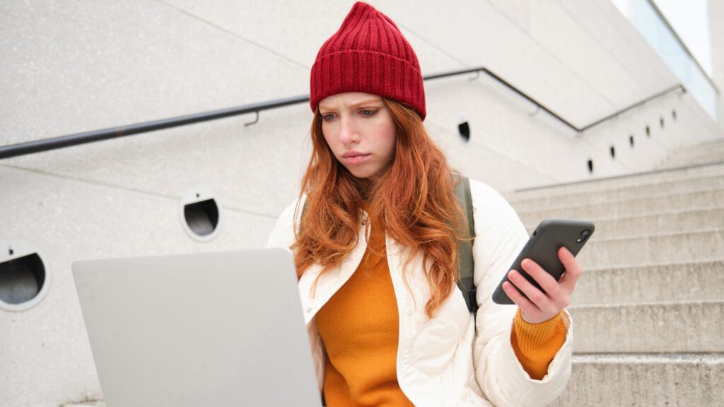 Portrait of redhead woman sits on stairs uses laptop and holds smartphone looks confused and upset