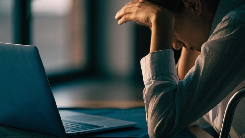 A person resting their forehead on one hand in front of a dark blue laptop on a dark desk mat