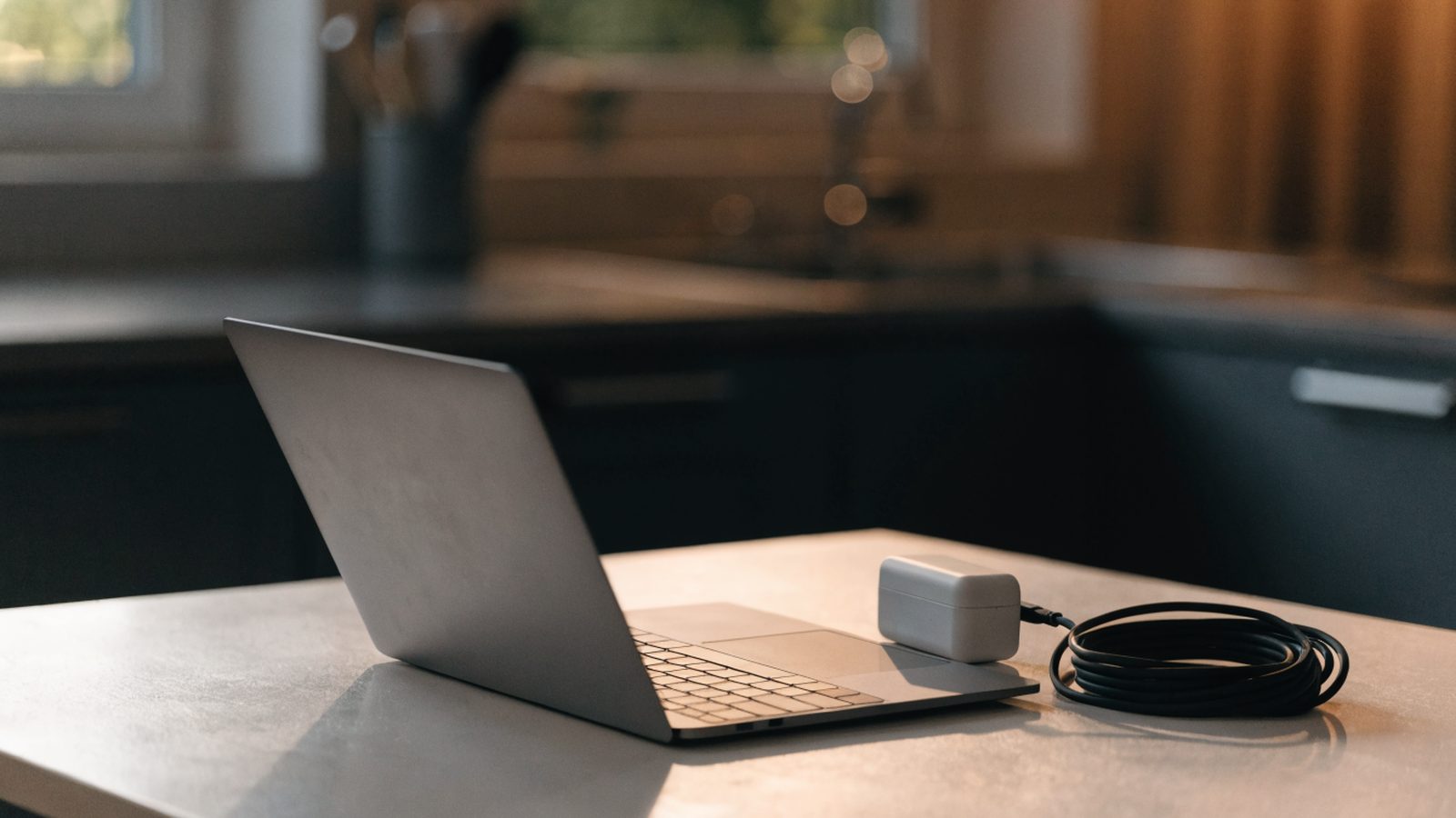 A matte grey laptop on a kitchen table next to a compact white power adapter with its cable neatly coiled