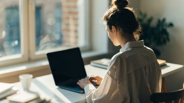 A person seen from behind working on a dark blue laptop in a bright home office