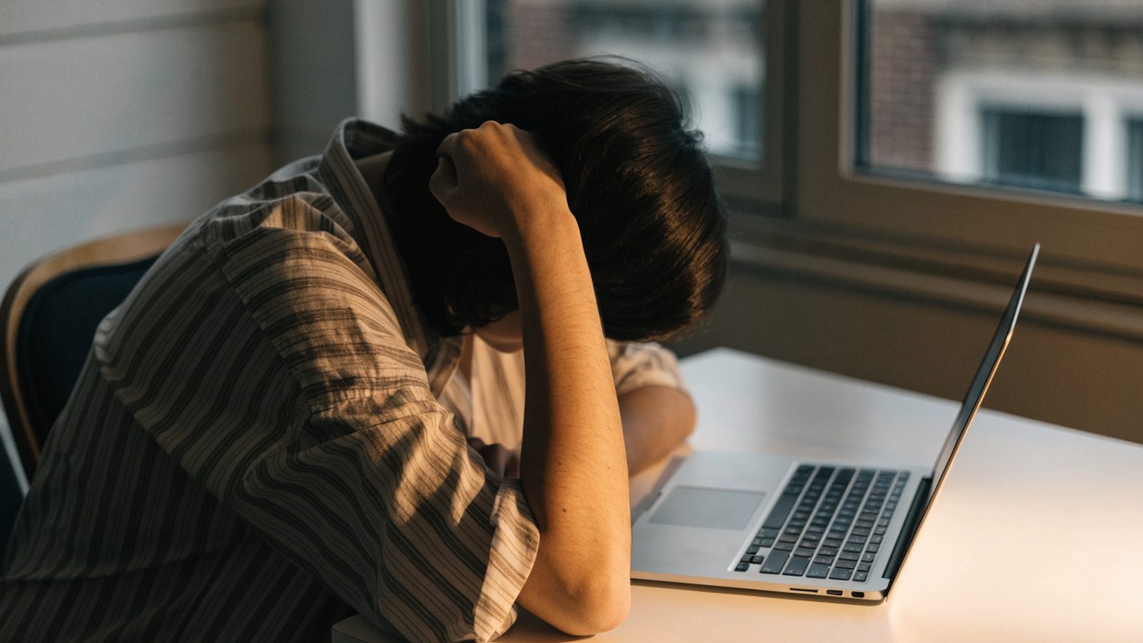 A person resting their forehead on one hand in front of a silver laptop on a white desk