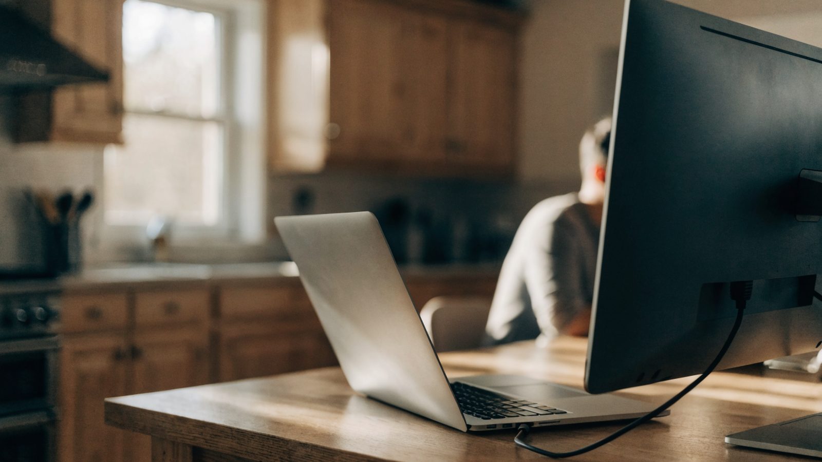 A silver laptop connected to a large external monitor with a blank dark screen via a cable on a kitchen table