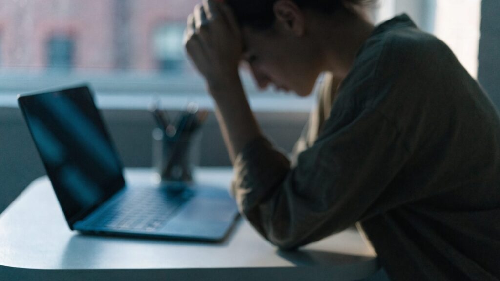 A person resting their forehead on one hand in front of a dark blue laptop on a white desk