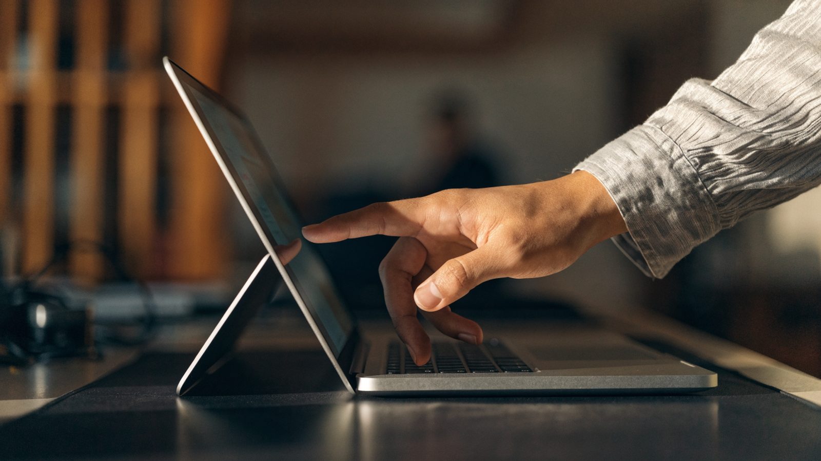 A hand touching the screen of a convertible silver laptop folded in tent mode on a dark desk mat