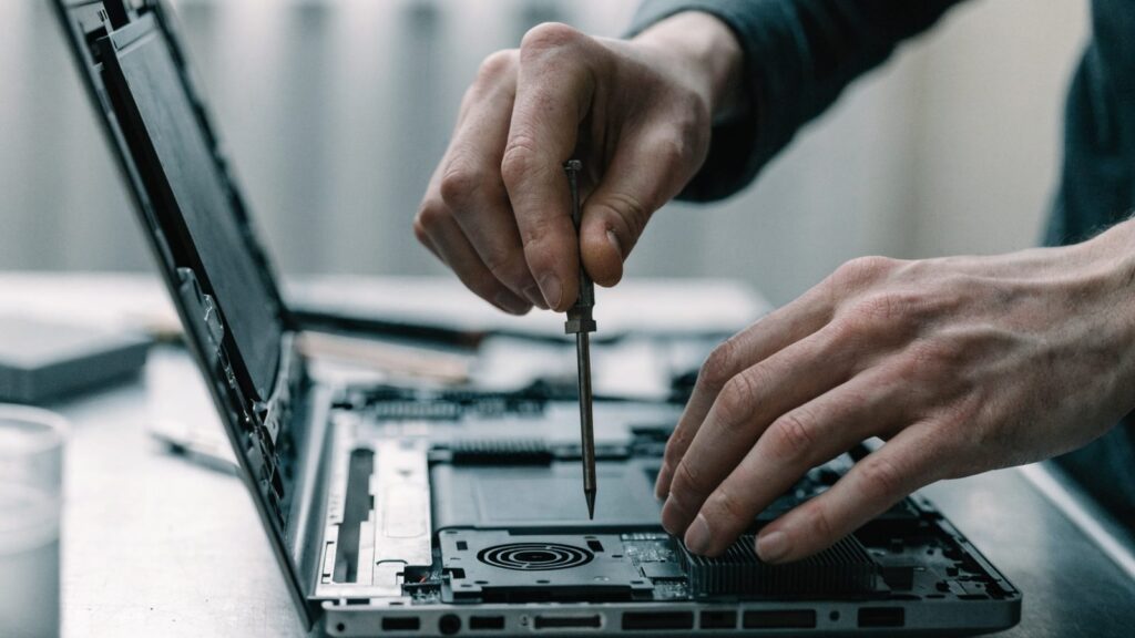 A technician's hands holding a small screwdriver over an opened graphite black laptop with internal components