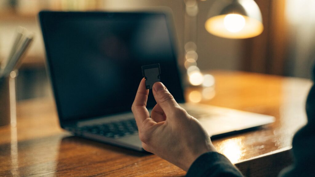 An SD card held between two fingers in front of a graphite black laptop on a wooden desk