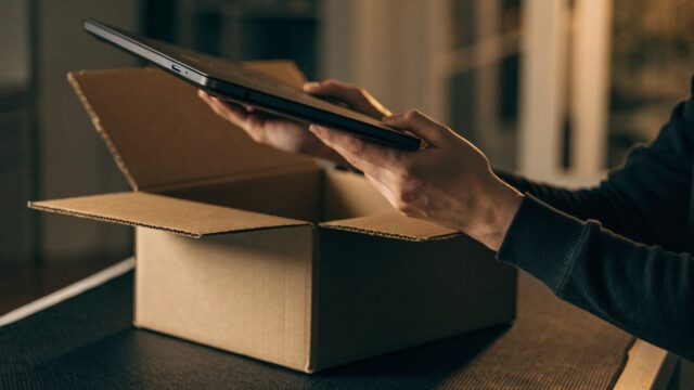 Hands lifting a brand-new graphite black laptop out of its plain cardboard box on a dark desk mat