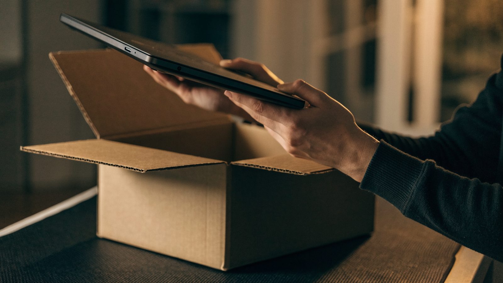Hands lifting a brand-new graphite black laptop out of its plain cardboard box on a dark desk mat