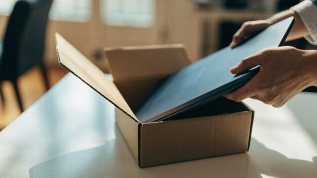 Hands lifting a brand-new dark blue laptop out of its plain cardboard box on a white desk