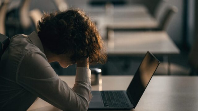 A person resting their forehead on one hand in front of a graphite black laptop on an office desk