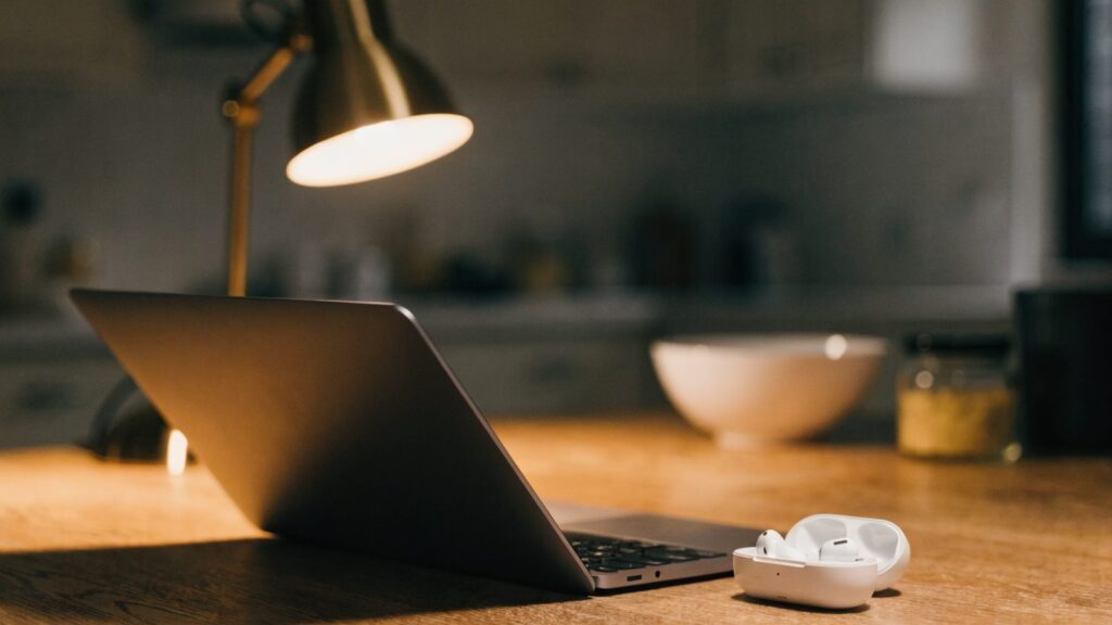 White wireless earbuds in their open case beside a matte grey laptop on a kitchen table
