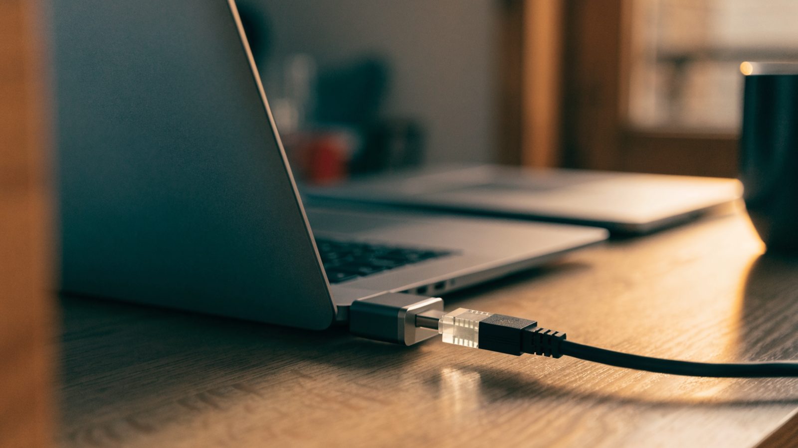 An ethernet cable being plugged into a USB-C adapter connected to a matte grey laptop on a wooden desk