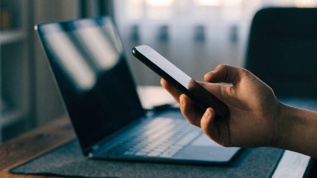 A hand holding a smartphone in front of a dark blue laptop on a dark desk mat