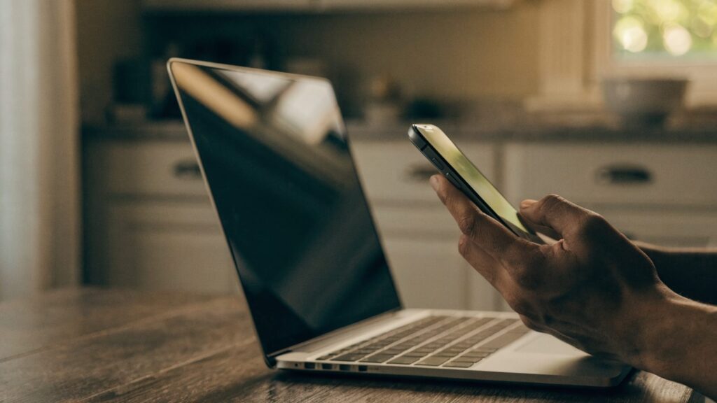 A hand holding a smartphone in front of a silver laptop on a kitchen table