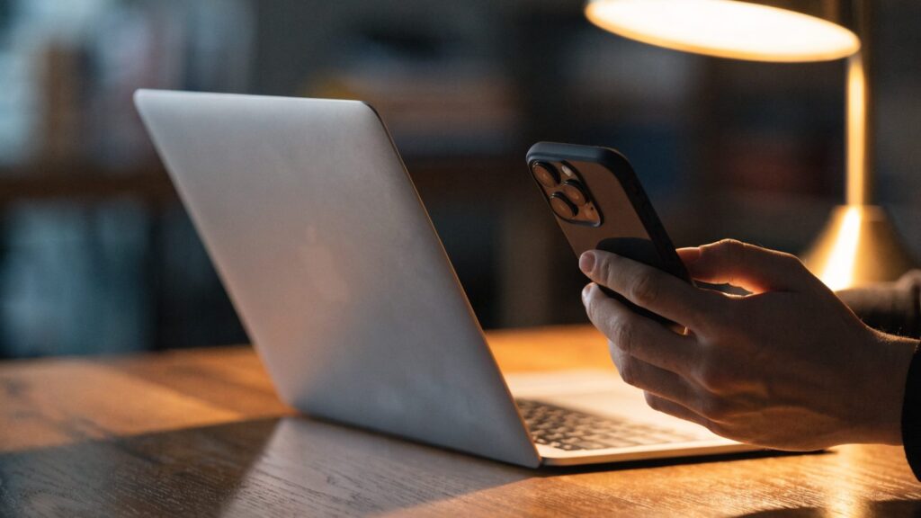 A hand holding a smartphone in front of a silver laptop on a wooden desk