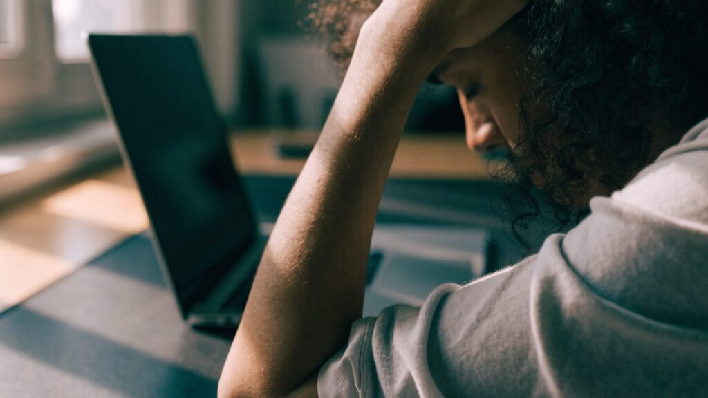 A person resting their forehead on one hand in front of a graphite black laptop on a dark desk mat