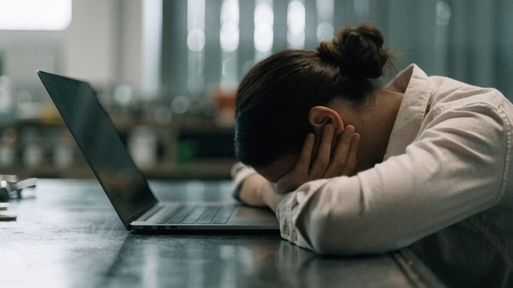 A person resting their forehead on one hand in front of a graphite black laptop on a workbench