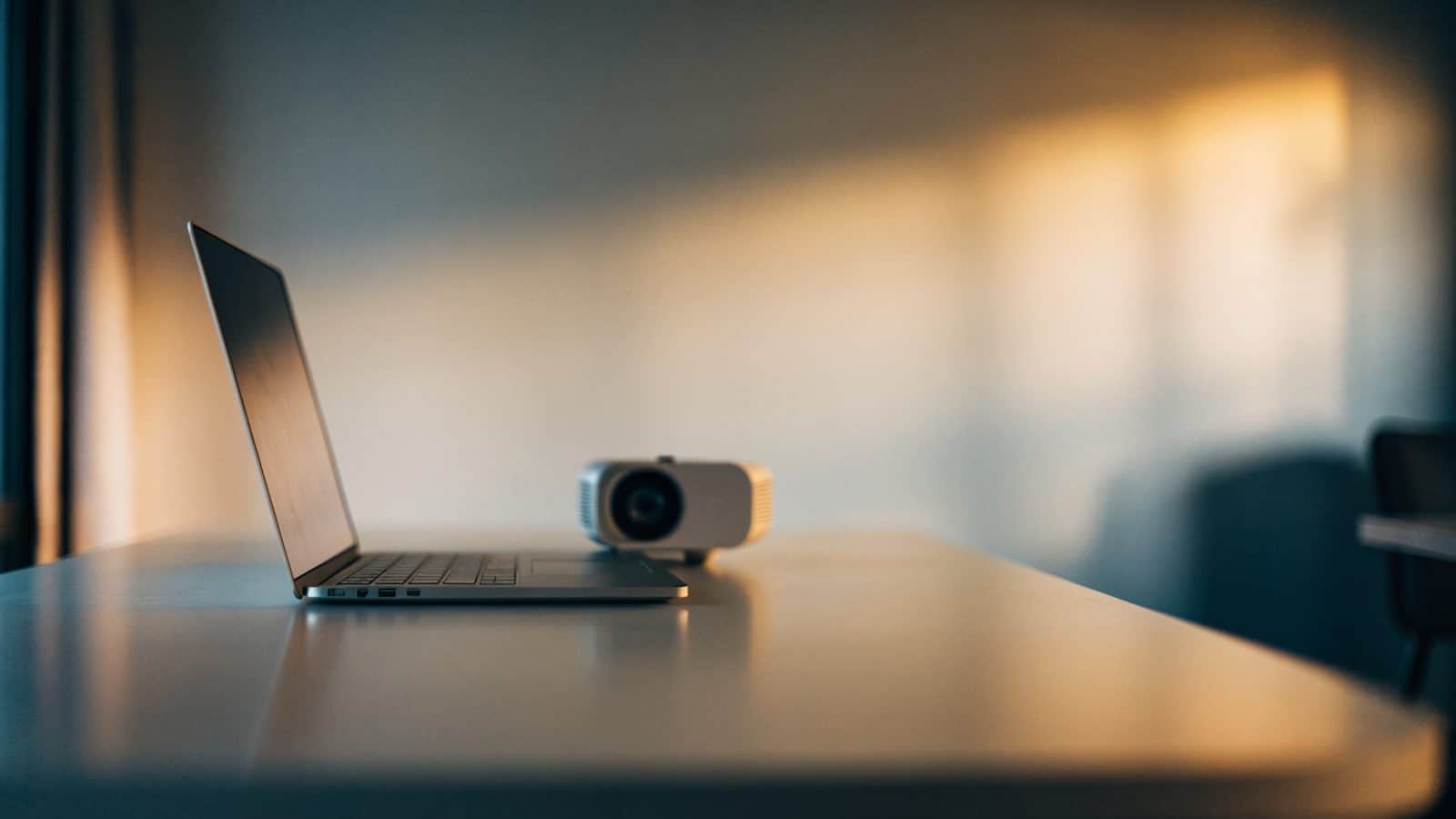 A silver laptop beside a small projector on a table