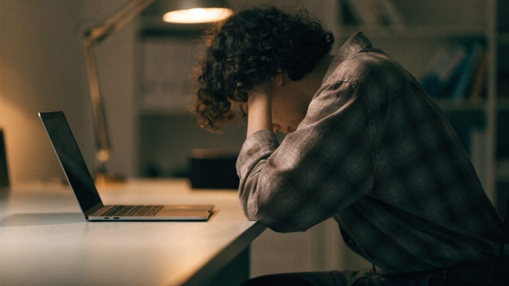 A person resting their forehead on one hand in front of a matte grey laptop on an office desk